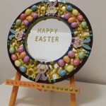 A round mirror on an eisel, decorated with easter eggs, happy easter is written on the mirror - recycled artwork by A round mirror on an eisel, decorated with easter eggs, happy easter is the message written on the mirror. Recyled artwork by Anne Bickley